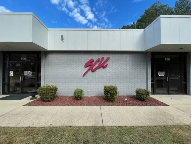 Exterior of a building with a pink SAL logo on a gray brick wall, landscaped with small bushes, two black doors, and a partly cloudy sky above.