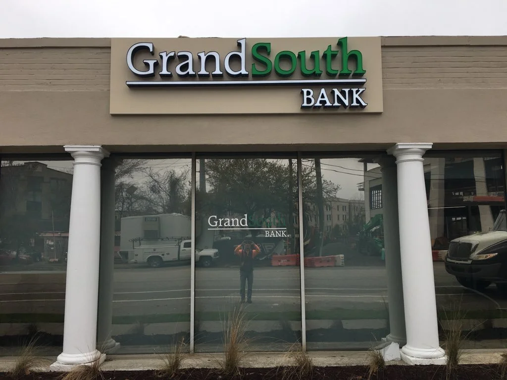 The storefront of Grand South Bank with a sign above the entrance. The sign features the bank's name in large letters, with "Grand" in black and "South" in green. The building has two white columns flanking glass doors, with a reflection of the stree