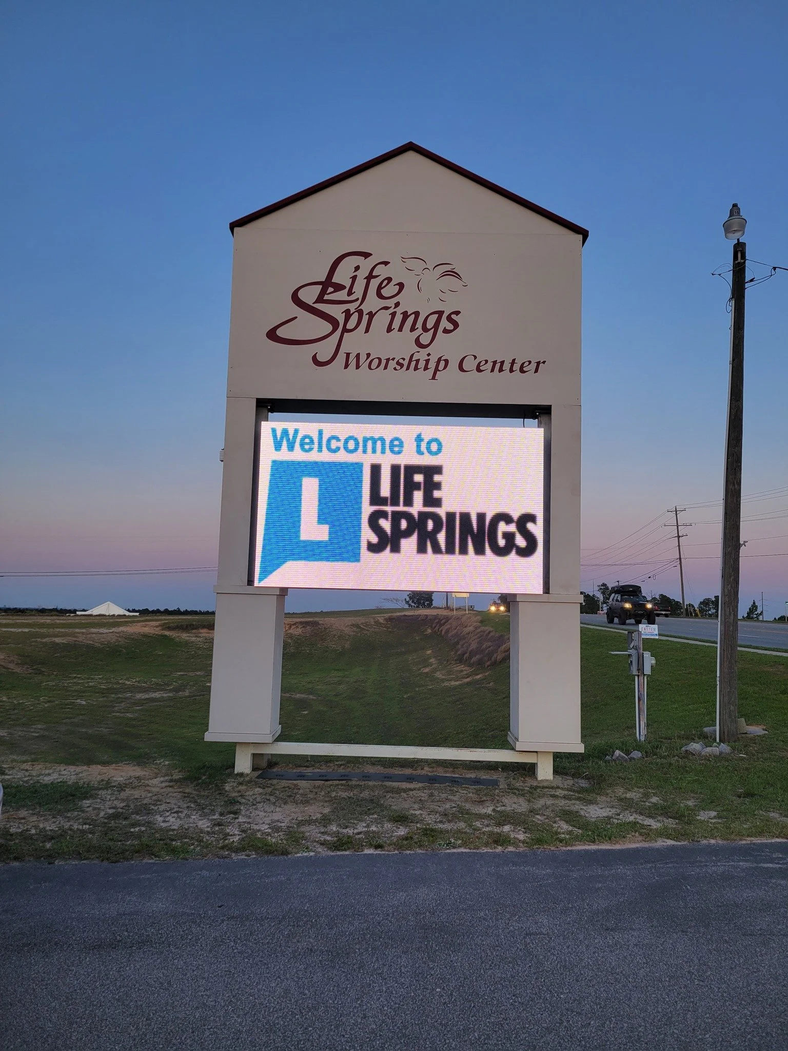 A large sign for Life Springs Worship Center, with a digital message board below it that says "Welcome to Life Springs." The sign is on the side of a road, with a grassy area and some vehicles in the background, under a clear sky during dusk.