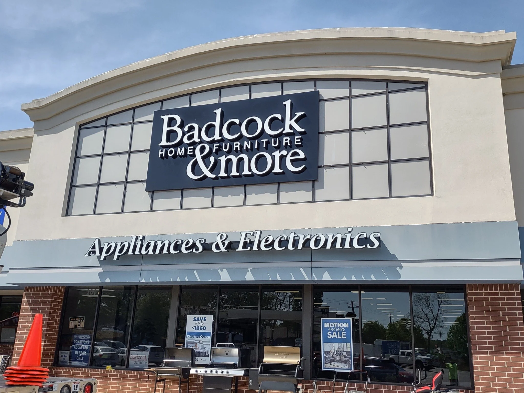 The exterior of a Baddock Home Furniture & More store with large windows, signage, and a brick facade, featuring a promotion for appliances and electronics.