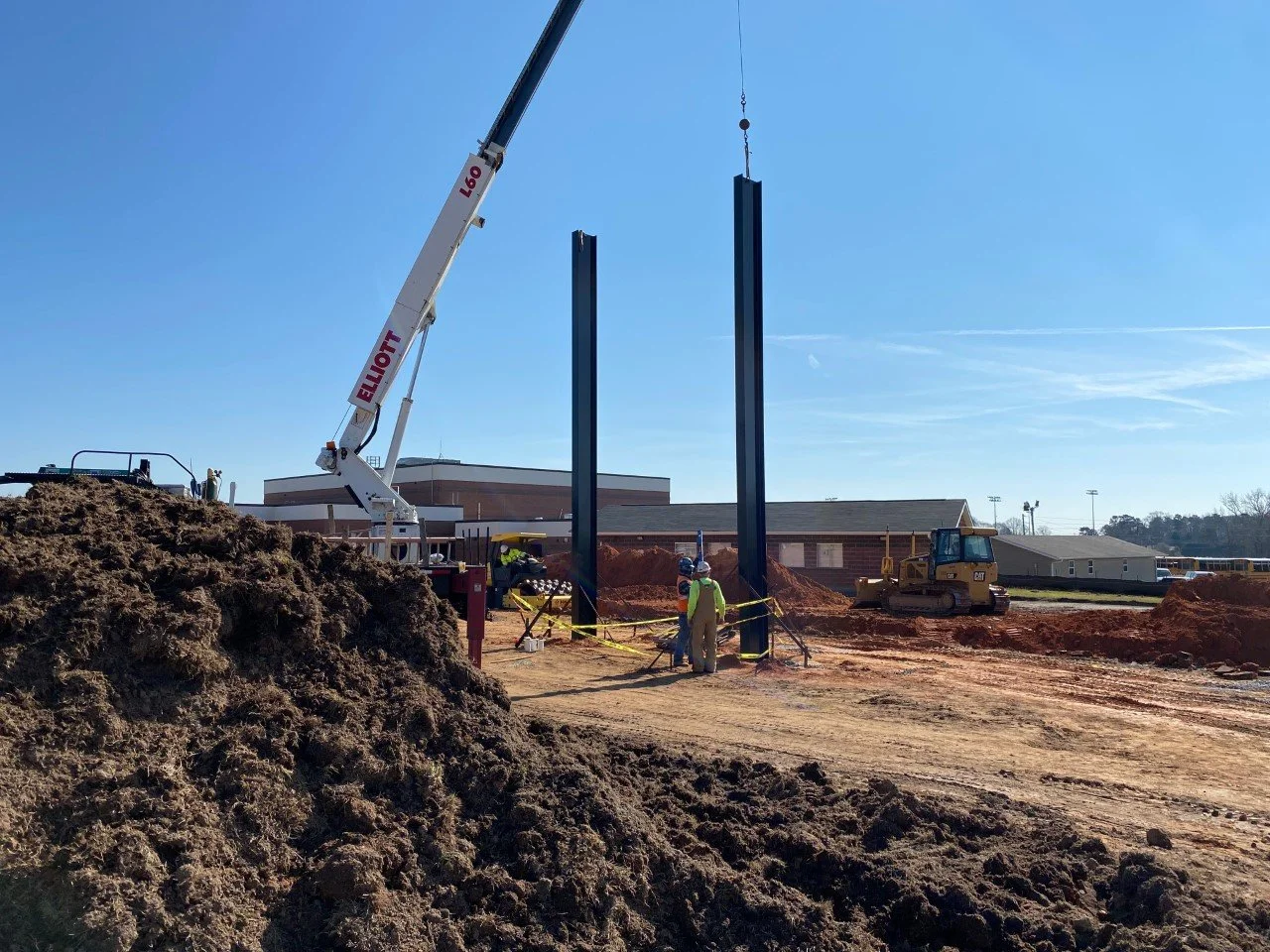 Construction site with a crane lifting steel beams and workers standing nearby, dirt mounds, and construction equipment.