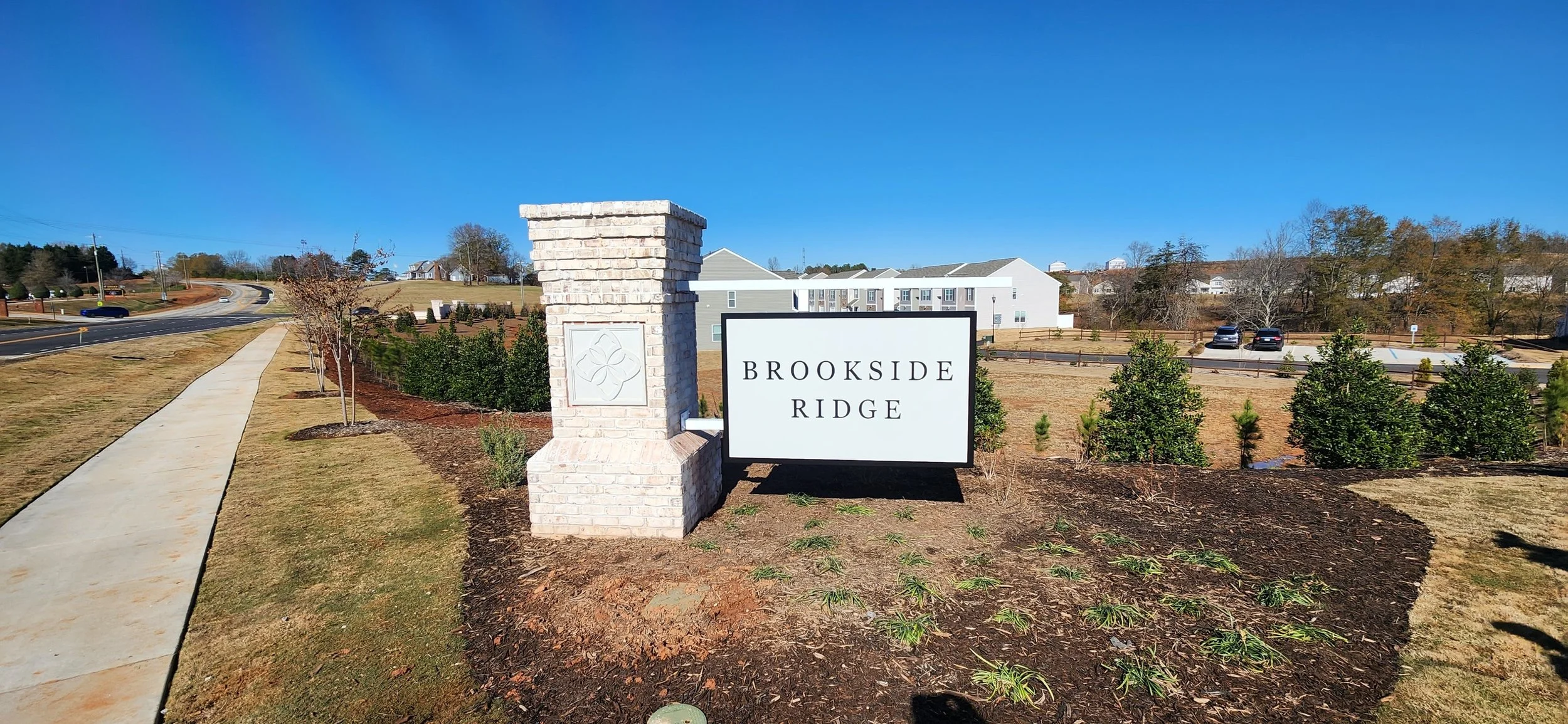 Entrance sign for Brookside Ridge residential community with a brick pillar and a white sign on landscaped ground, with a sidewalk and parking lot in the background under a clear blue sky.
