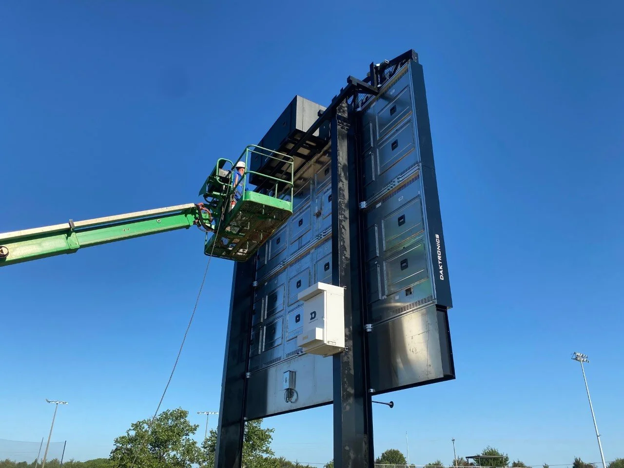 Worker on a green aerial lift inspecting or maintaining a tall electronic scoreboard at a sports field on a clear day.
