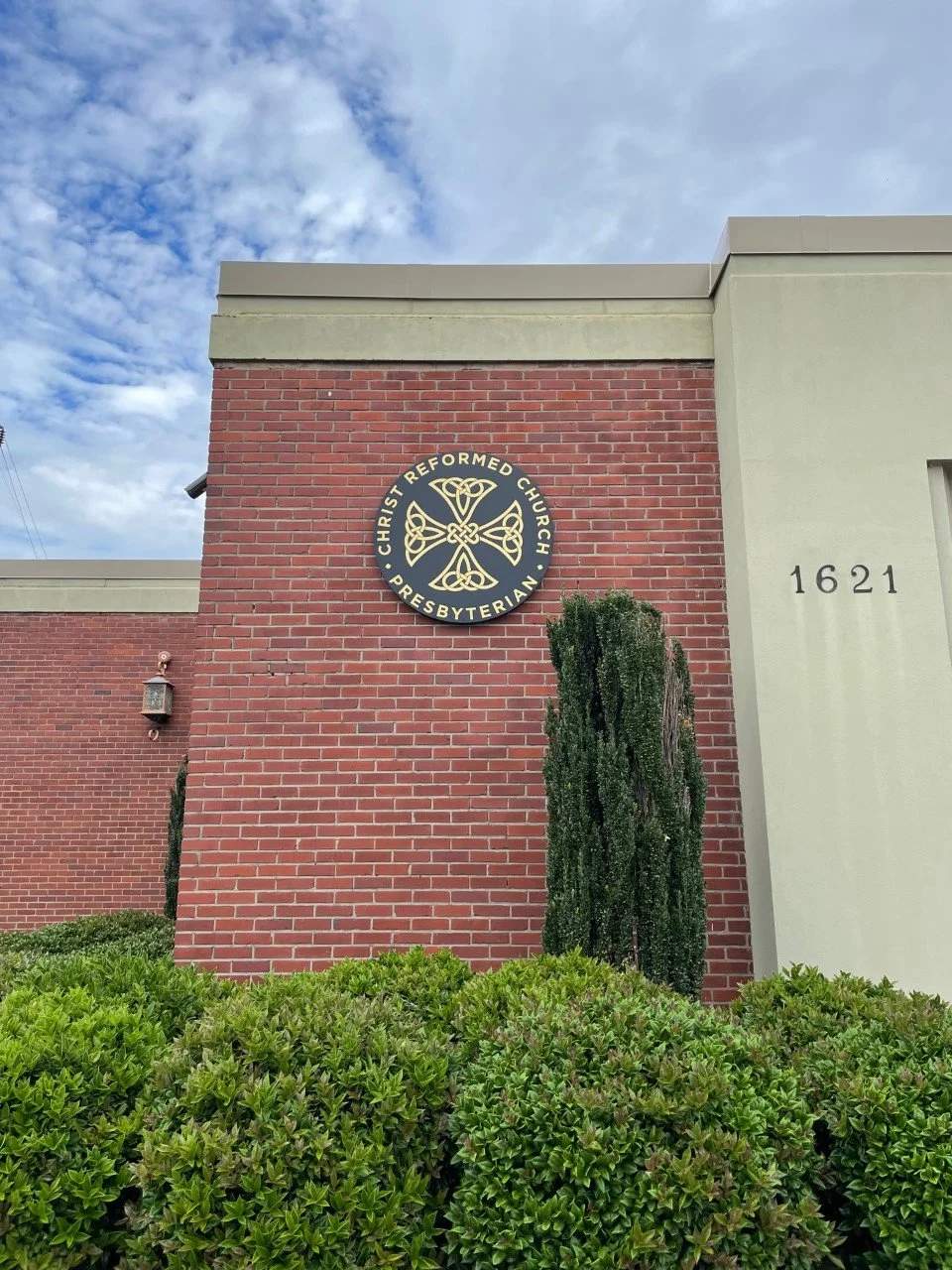 Exterior view of a church building with a brick and white facade, a sign reading 'Christ Reformed Church Presbyterian,' and the number 1621 on the wall. There are bushes and a tall, narrow tree in front, with a partly cloudy sky overhead.