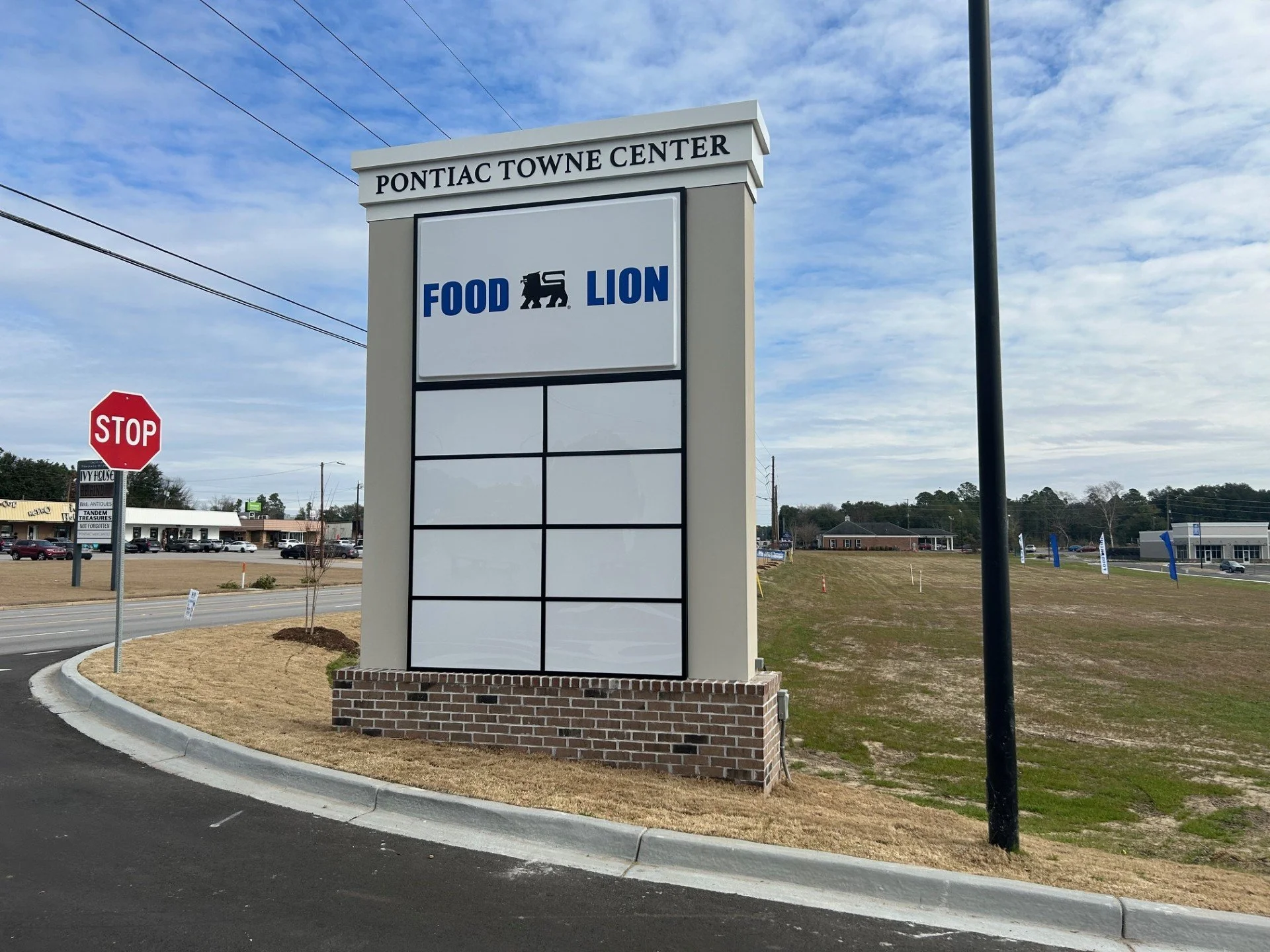 The image shows a large sign for the Pontiac Towne Center, with a smaller sign in the background that reads 'STOP' in red, along with various shops and parked cars in a shopping plaza area under a partly cloudy sky.
