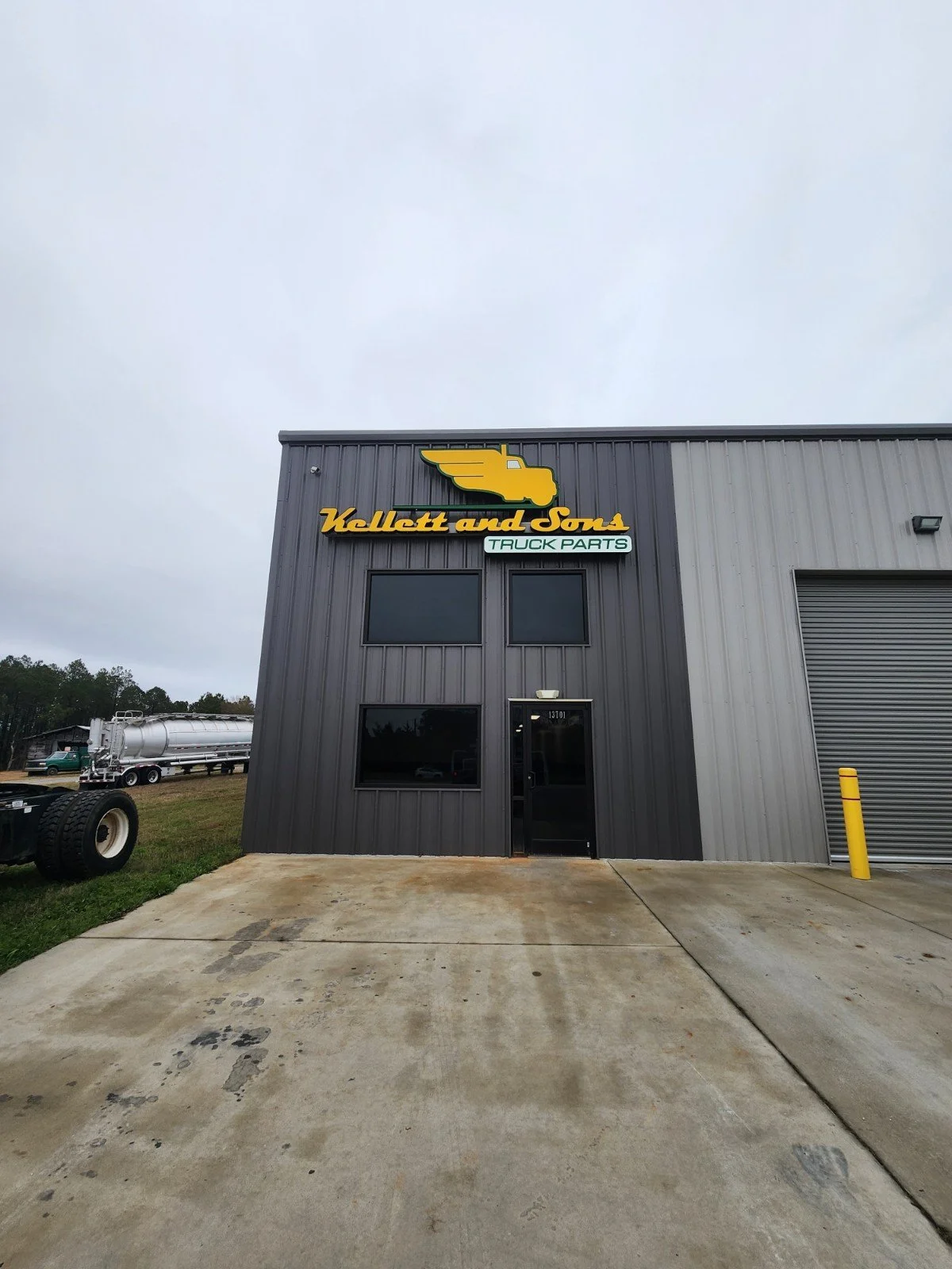 Exterior of a gray industrial building with a yellow and black logo reading 'Kellert and Sons Truck Parts'. The building has three windows, a black door numbered 13101, and a large garage door. There is a parking lot and a propane truck visible in th