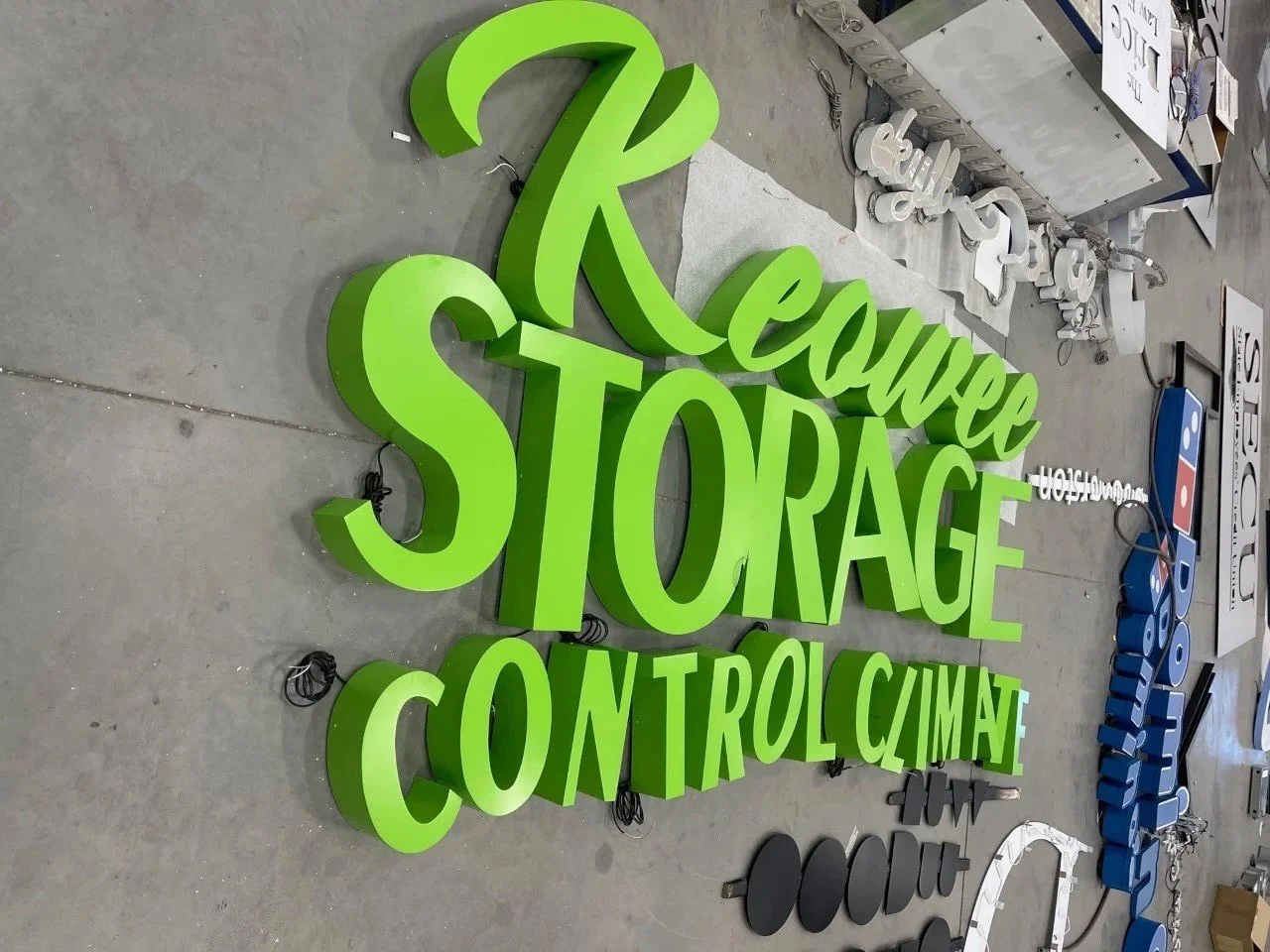 A large, green, three-dimensional sign with the words 'Recycle,' 'STORAGE,' and 'CONTROL CLIMATE' lying on a concrete floor, with other signs and materials nearby.