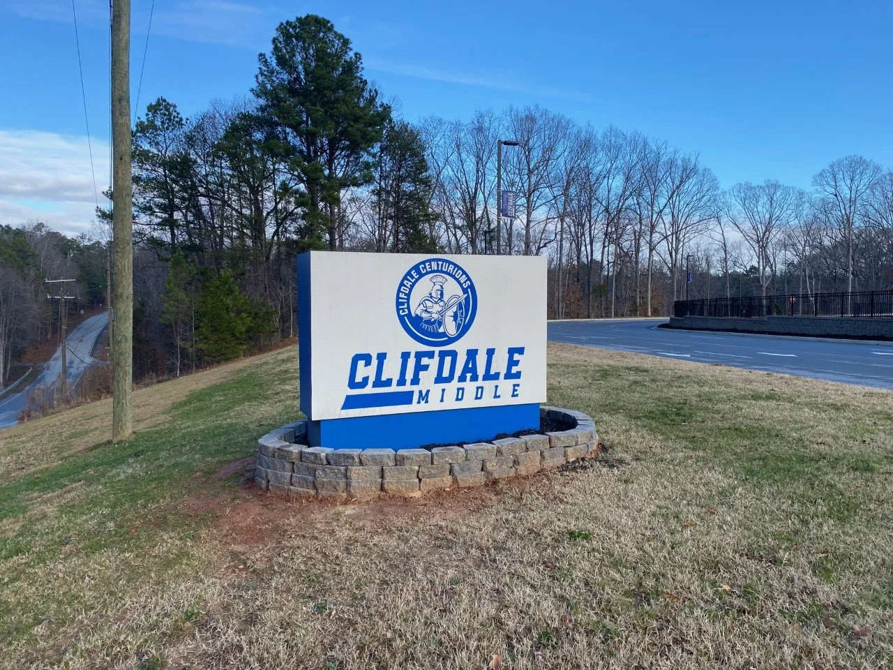 A sign for Clifdale Middle School with a logo of a Spartan helmet and shield, located on a grassy area near a road, with trees and a blue sky in the background.