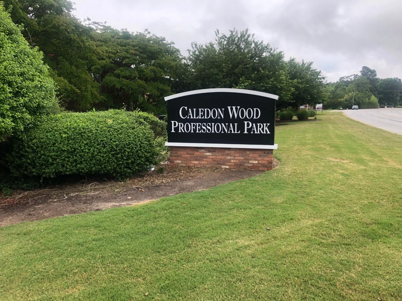 Sign for Caledon Wood Professional Park next to a grassy area and sidewalk, with trees and shrubs in the background.