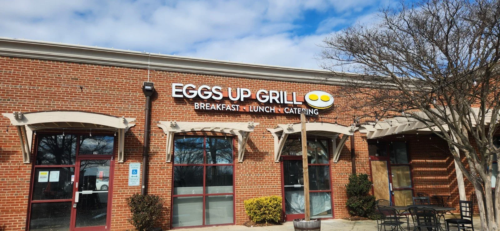 A brick storefront with a sign that reads 'Eggs Up Grill' and indicates breakfast, lunch, and catering. There are large windows, some with curtains, and an outdoor seating area with chairs and a tree without leaves.