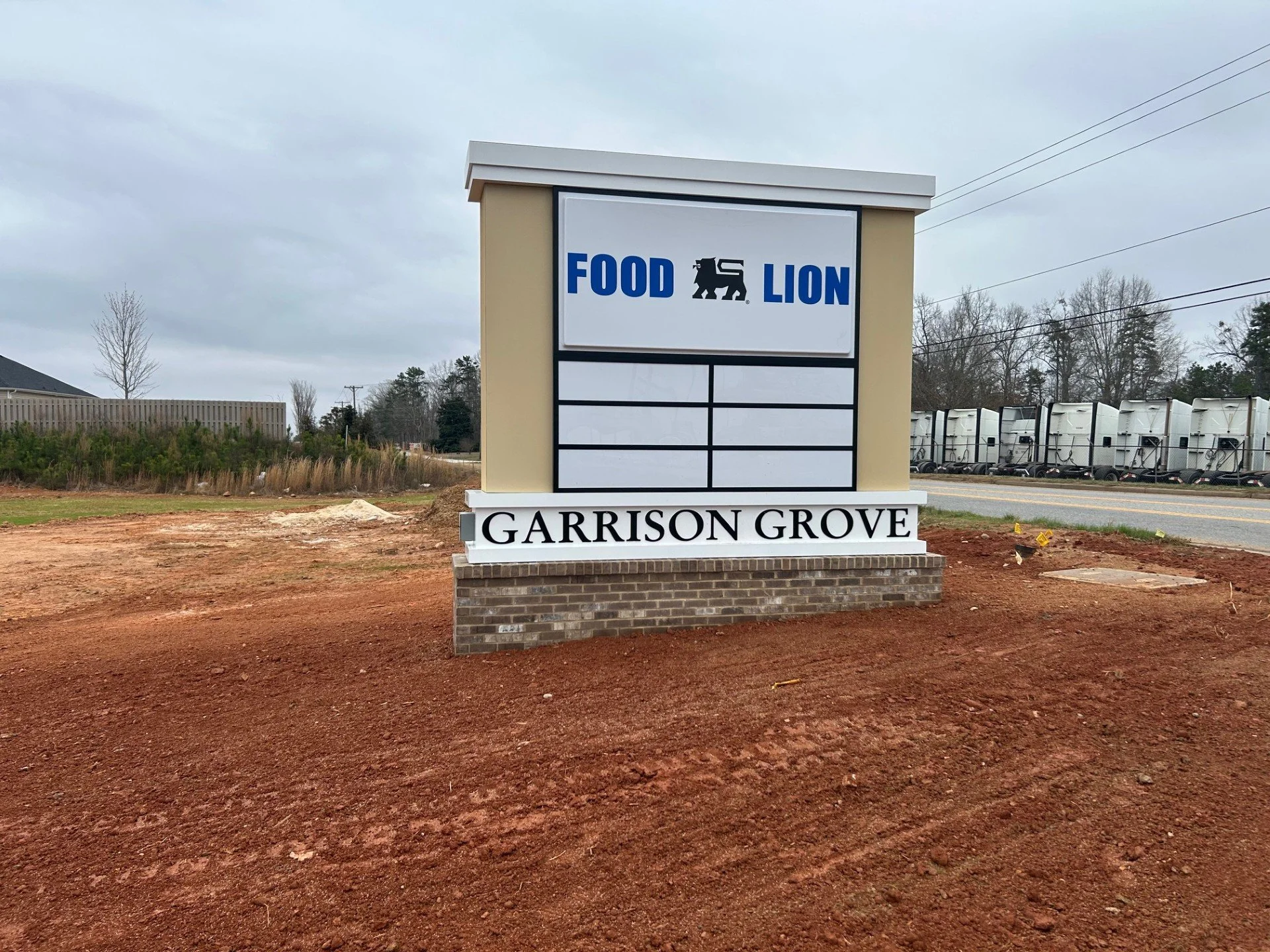 A roadside sign for Garrison Grove with a logo for Food Lion in the upper part. The sign has an empty area for additional information and the background shows a cloudy sky, trees, and a row of trailers.