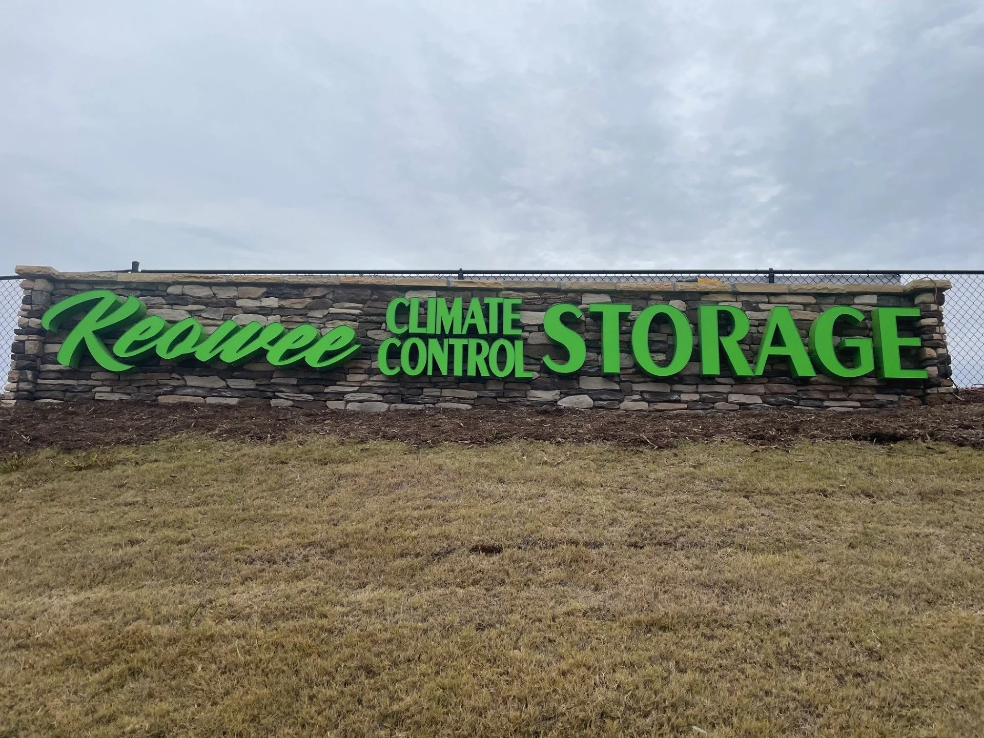 A stone sign with green illuminated text that reads 'Reeves Climate Control Storage' against a cloudy sky, with a grassy area in the foreground.
