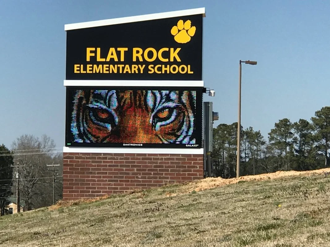 School sign for Flat Rock Elementary School with yellow text, tiger face digital display, and a paw print logo against a blue sky background.