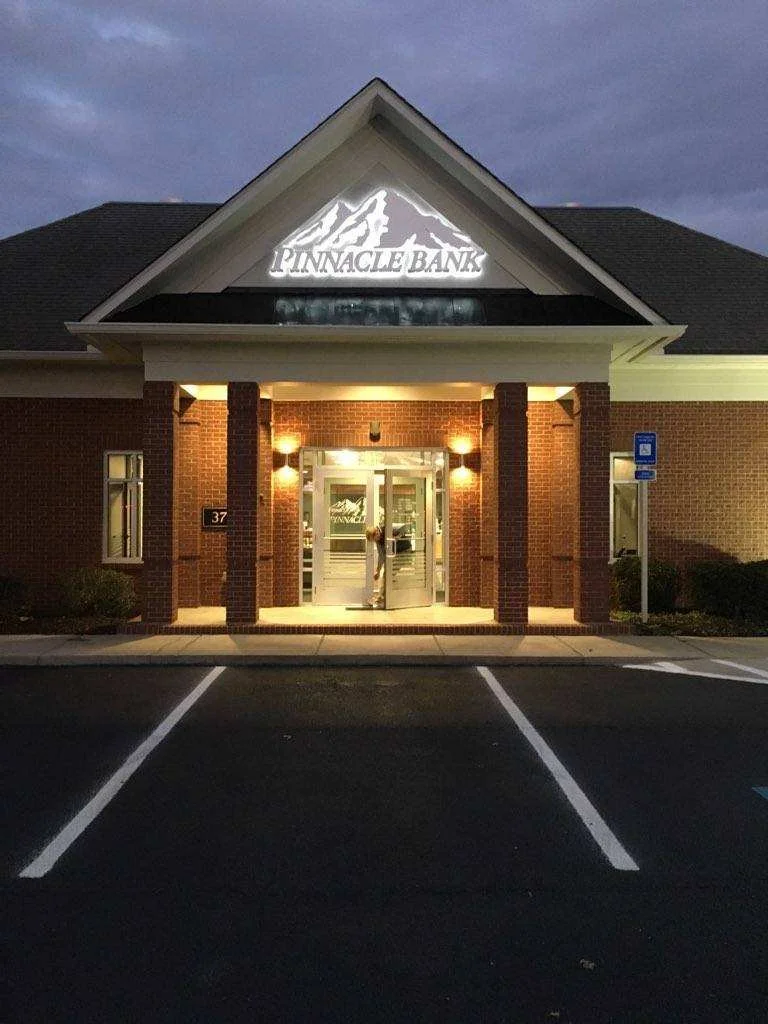 Exterior of Pinnacle Bank branch at night with illuminated sign, brick facade, and a person entering through glass doors. Parking lot in foreground with designated accessible parking space.