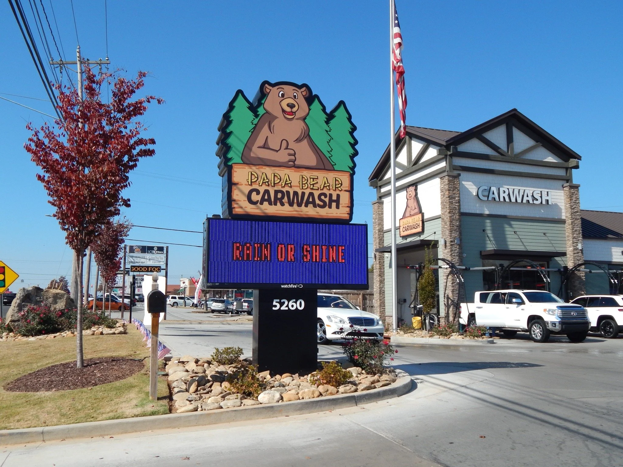Sign for Papa Bear Carwash with a cartoon bear giving a thumbs up, a flagpole with the American flag, a building with carwash signage, and a parking lot with various cars.