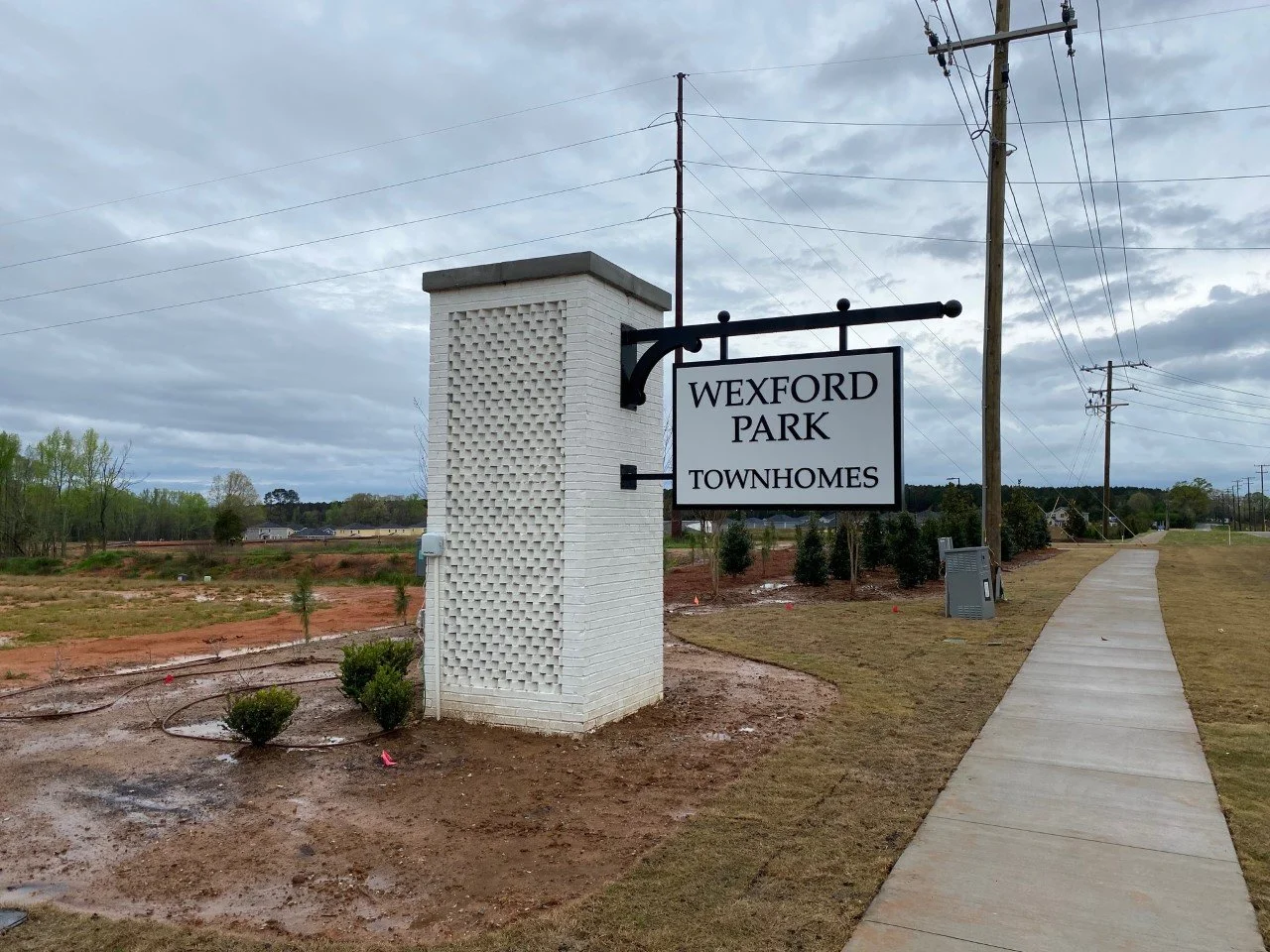Sign indicating Wexford Park Townhomes next to a sidewalk with utility poles and a mostly cloudy sky.
