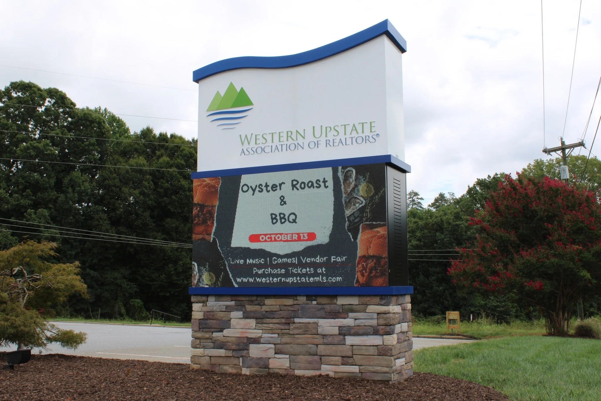 Large outdoor sign for Western Upstate Association of REALTORS with green mountain and blue wave logo at the top, and advertising for an oyster roast and BBQ event on October 15, with live music, vendor fair, and ticket purchase information.