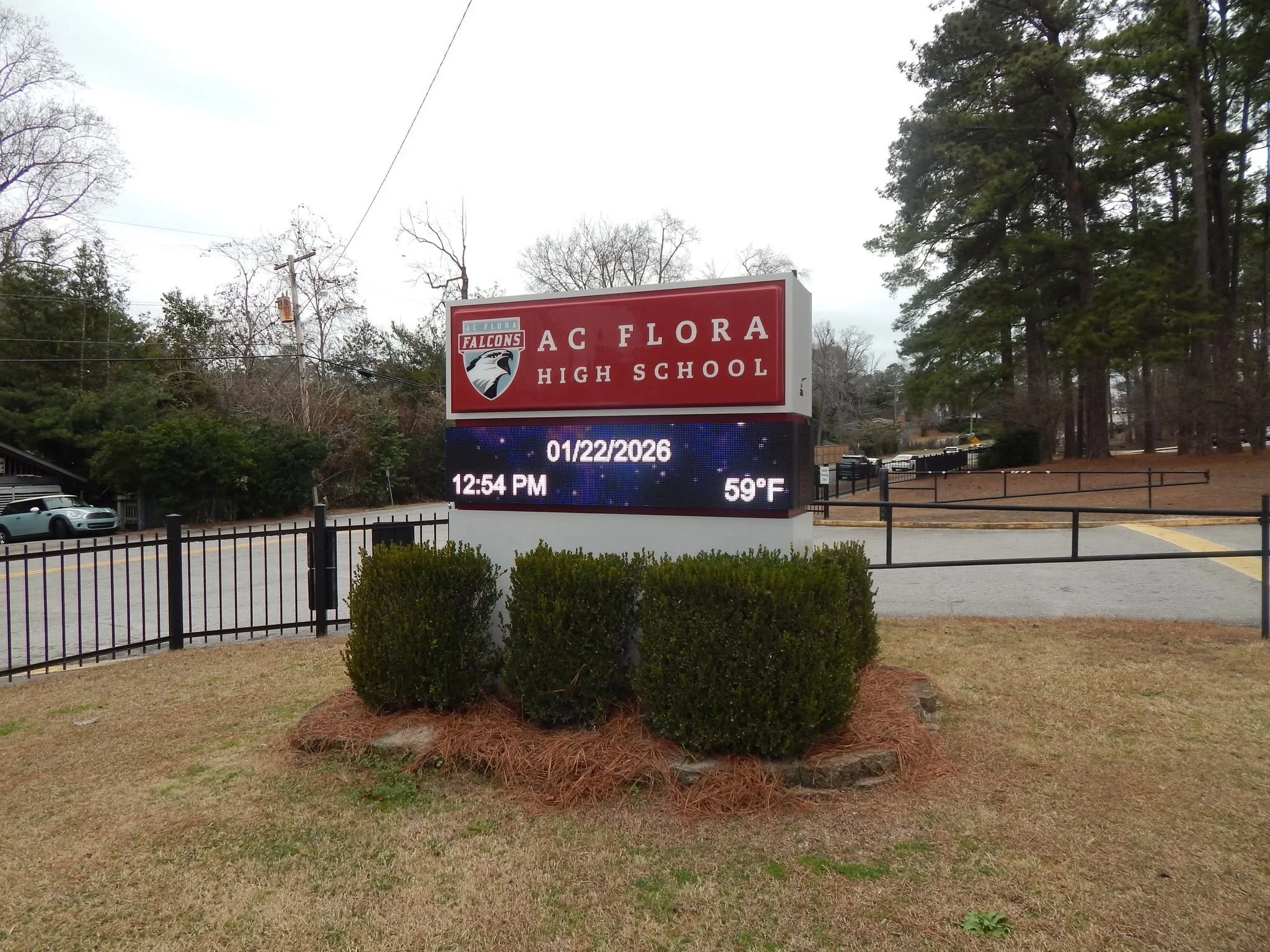 Signboard for AC Flora High School with a Falcon logo, digital display showing date, time, and temperature, surrounding trees, parked cars, and a black fence.