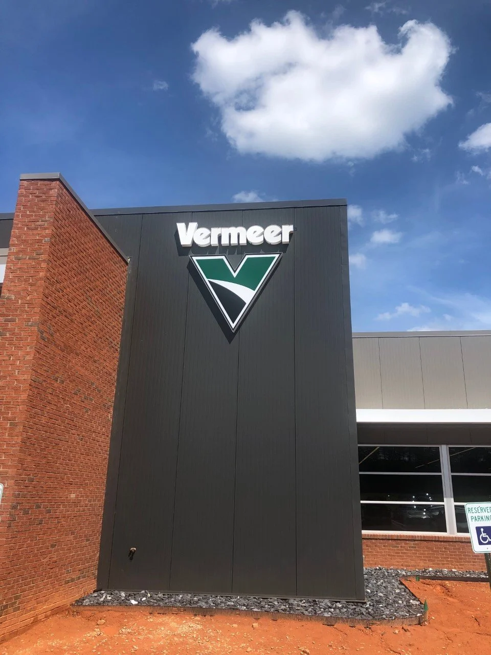 Exterior of Vermeer building with a large black sign featuring the Vermeer logo and name, brick and metal walls, a reserved parking sign, and a cloudy blue sky.