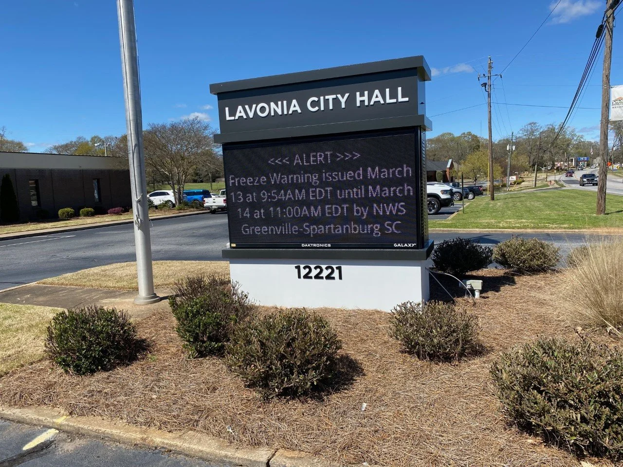 A digital sign outside Lavonia City Hall displaying a weather alert about a freeze warning issued in March, located on a patch of landscaped grass with shrubs, near a road with cars and power lines.