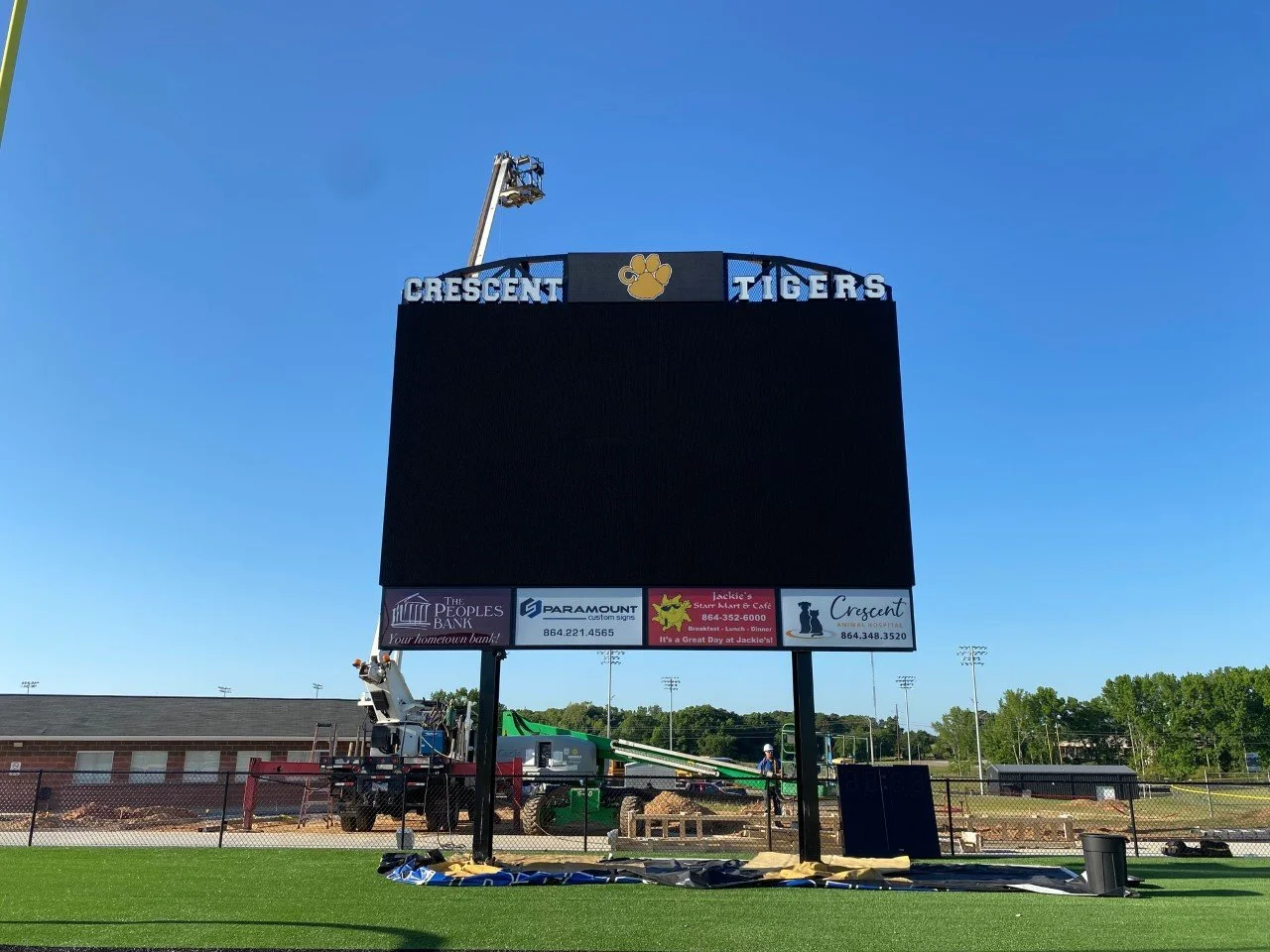 Large, empty digital scoreboard on a sports field with Crescent Tigers at the top and a paw print logo in the center. Construction equipment and workers are seen below the scoreboard, with a clear blue sky above.
