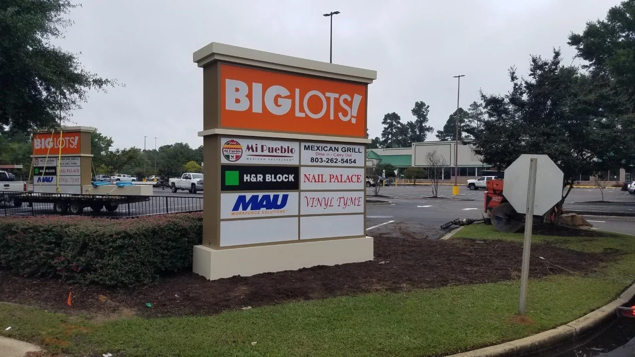 A large sign in a parking lot with the text 'BIG LOTS!' at the top, listing various stores including Mi Pueblo, Mexican Grill, Nail Palace, H&R Block, MAU workforce solutions, and Vinyl Time.