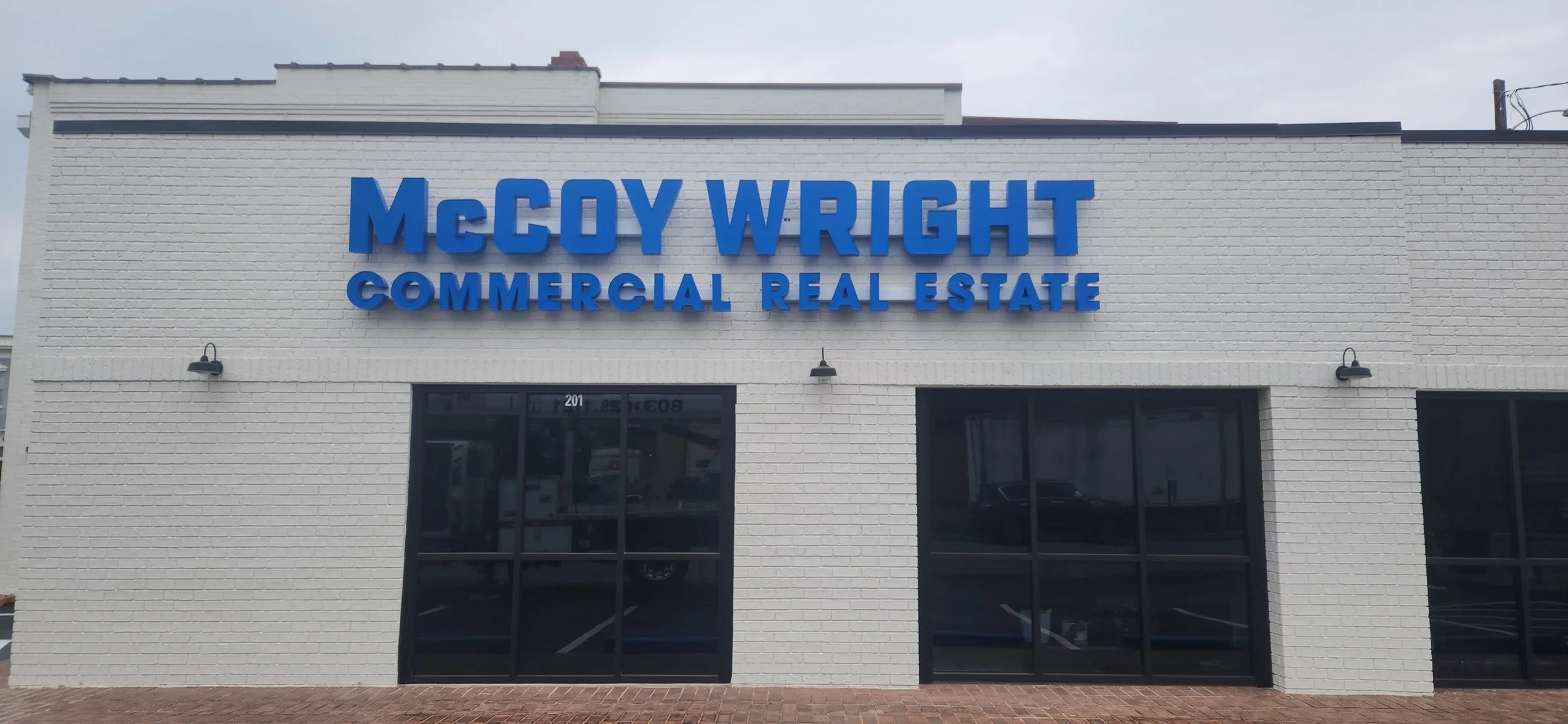 The front of a white brick building with blue signage reading 'McCoy Wright Commercial Real Estate' and two black exterior lights above the windows.