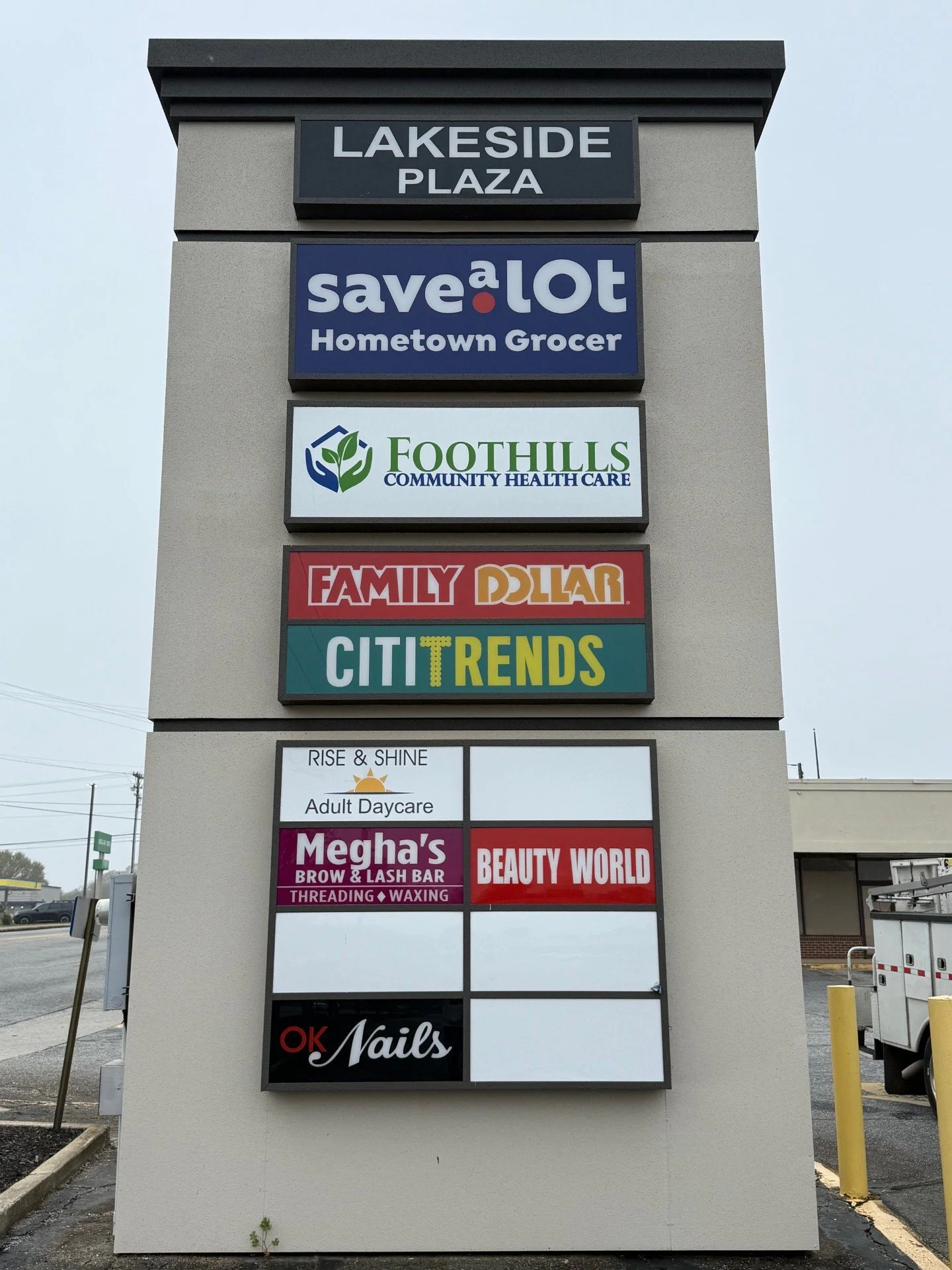 Signboard with multiple business signs including Lakeside Plaza, Save a Lot Hometown Grocer, Foothills Community Healthcare, Family Dollar, CitiTrends, Rise & Shine Adult Daycare, Megha's Beauty World, and OK Nails.