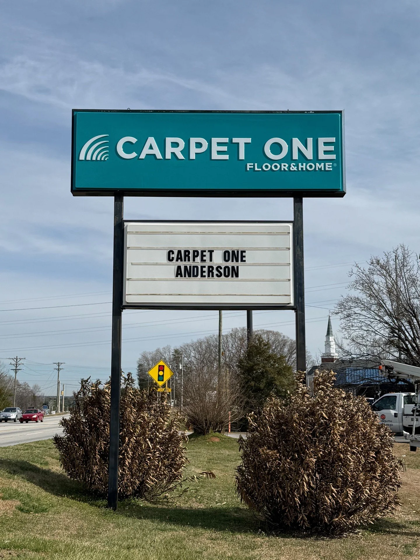 Sign for Carpet One Floor & Home with the words 'Carpet One Anderson' on a white background, set in a landscaped area with bushes and a road with cars and traffic lights in the background.