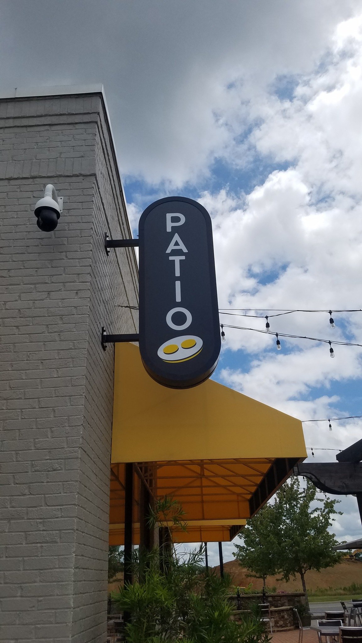 Business signage for a patio with a blue oval sign that reads 'Patio' with images of two yellow coins at the bottom, hanging outside a building with a brick wall and yellow awning, under a daytime cloudy sky.