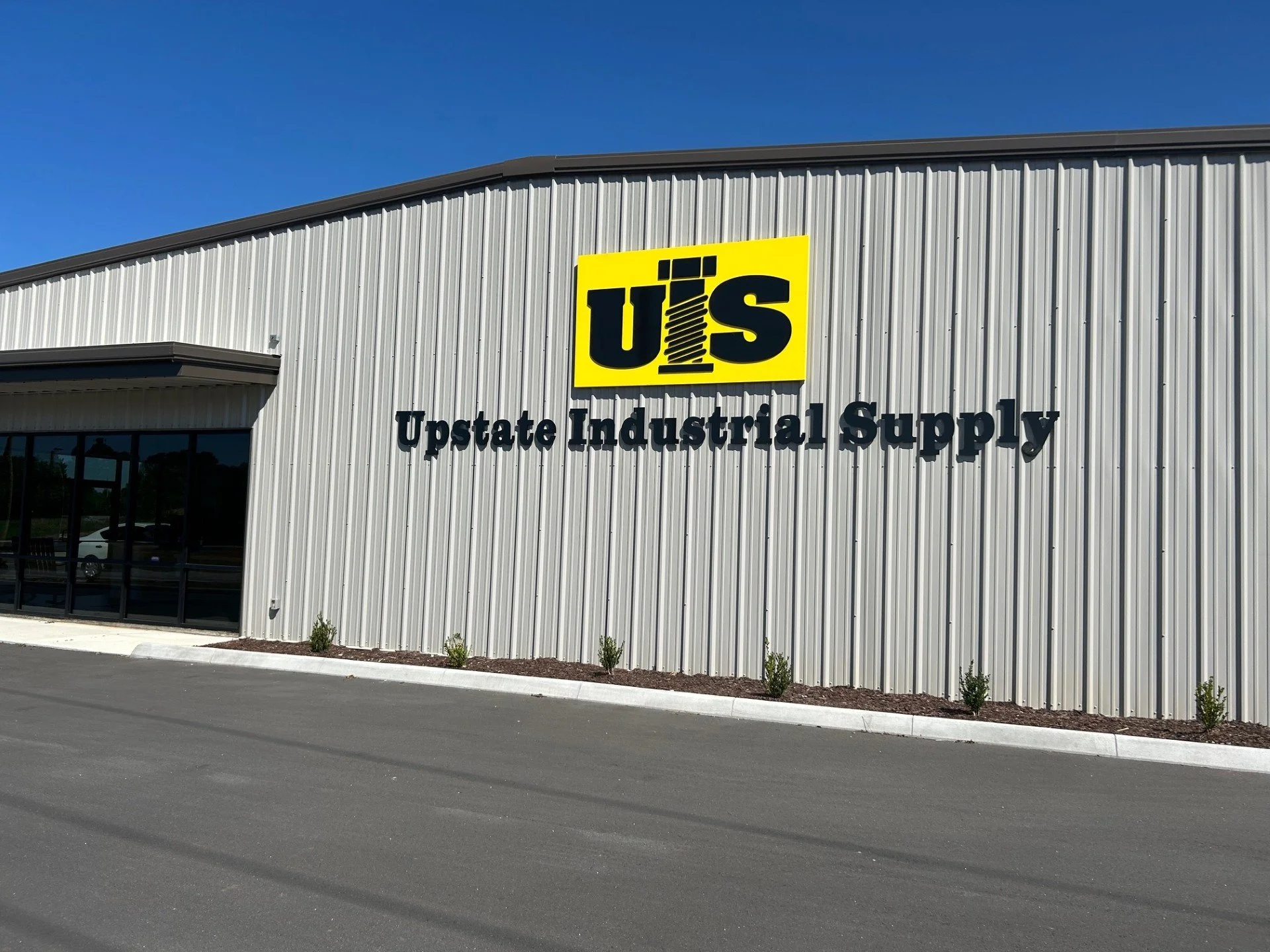 The exterior of a building with a large sign that reads 'U.S. Upstate Industrial Supply'. The building has silver metal siding and a row of small plants along the sidewalk in front.