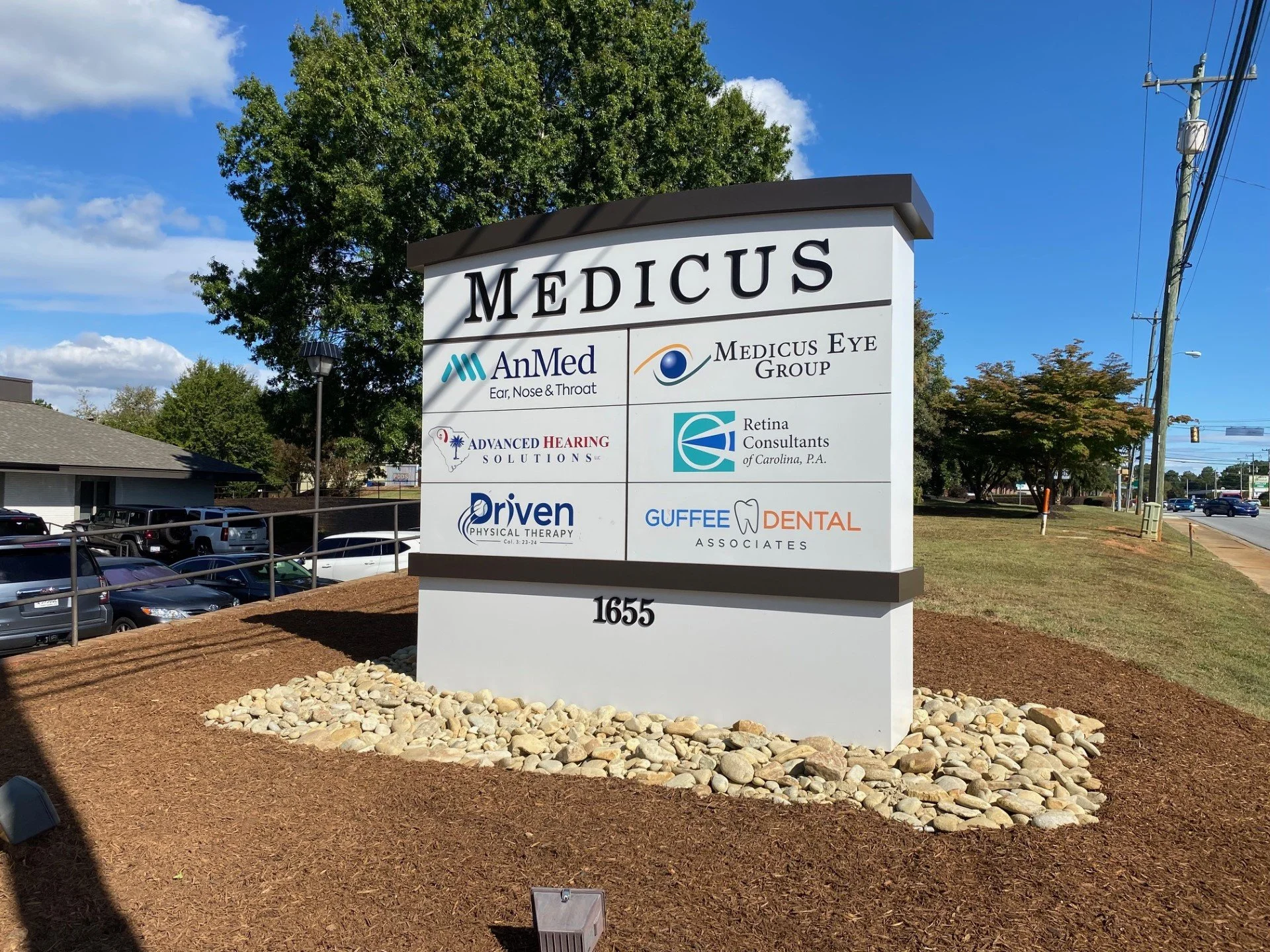 A tall outdoor sign for Medicus with logos of healthcare providers, including AnMed, Medicus Eye Group, Advanced Hearing Solutions, Retina Consultants, Driven Physical Therapy, Guffee Dental, and others, outside a medical complex on a clear sunny day