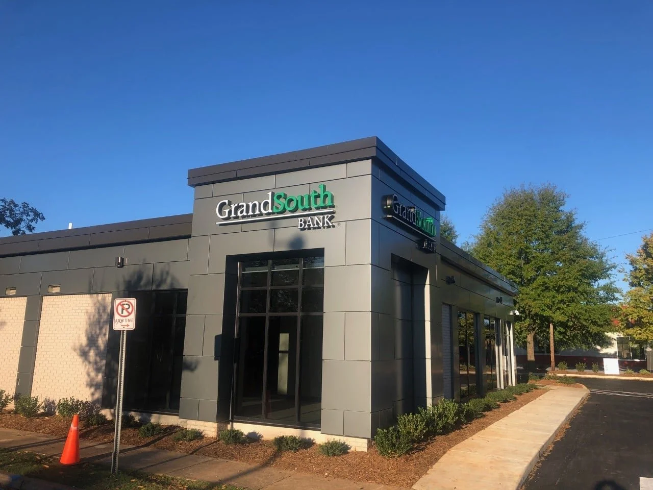 Exterior of a gray bank building with a sign reading 'GrandSouth Bank' and a sidewalk in front, surrounded by small bushes and a no parking sign.