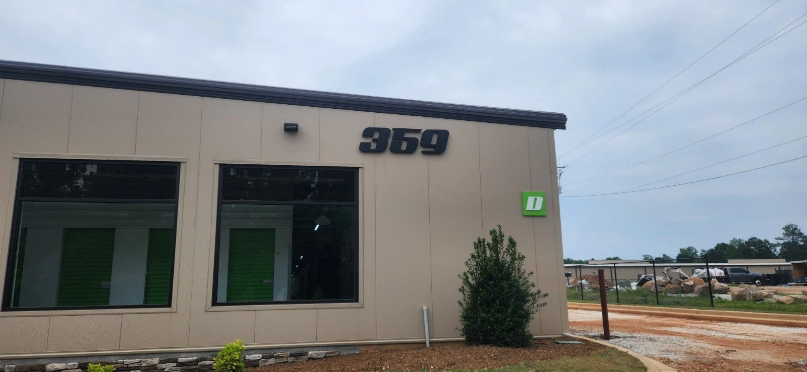 Exterior of a commercial building with the number 359, large windows with green window coverings, a small shrub, and a green Dollar General sign.
