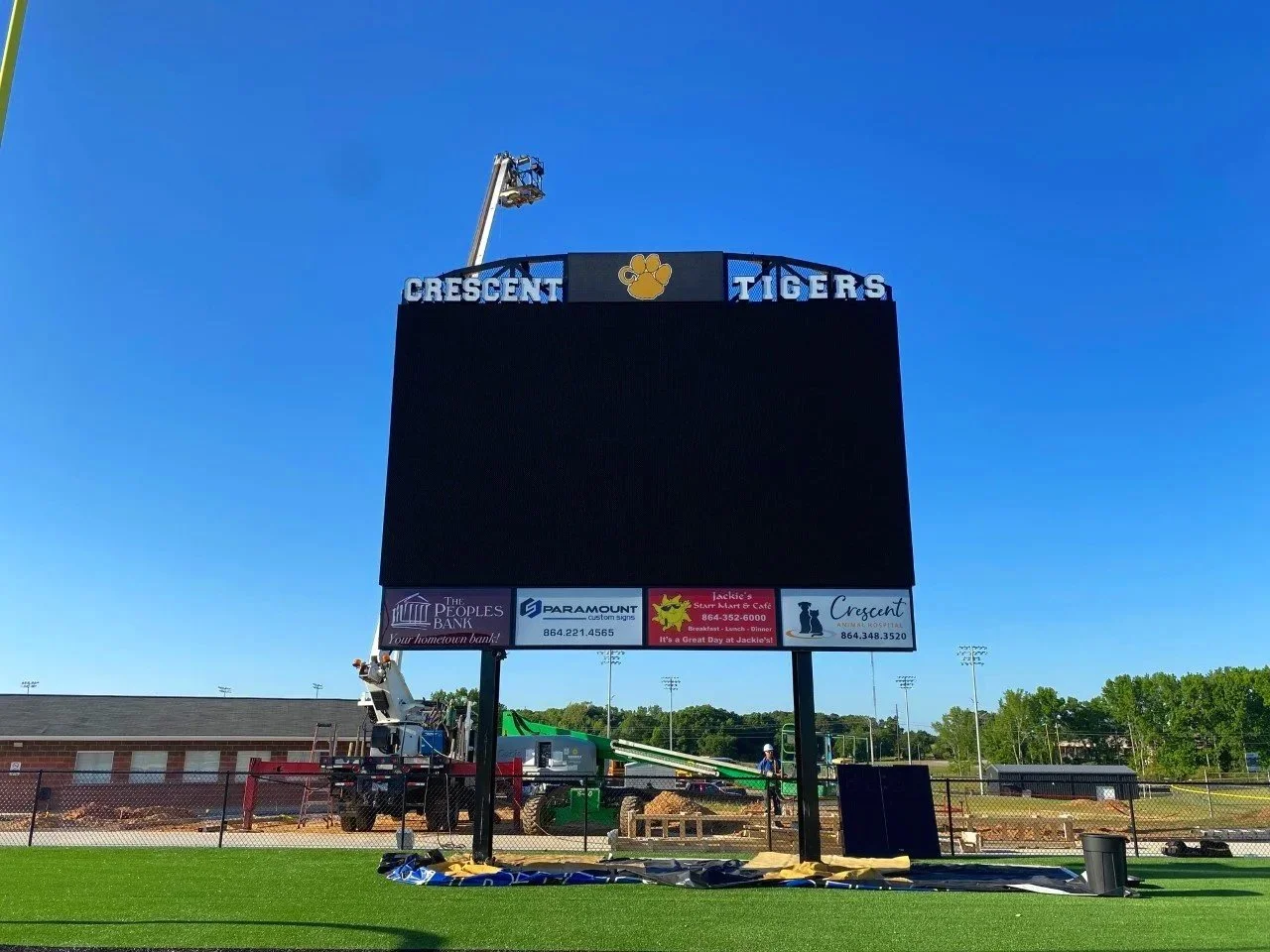 A large electronic scoreboard at a sports field being built, with a crane lifting equipment near the top, blue sky, and a few trees in the background.
