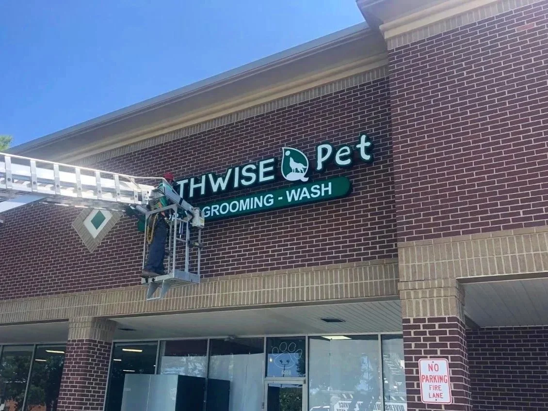 A worker on a lift installing or repairing a sign for 'Thwise Pet' offering grooming and wash services on the brick exterior of a store.