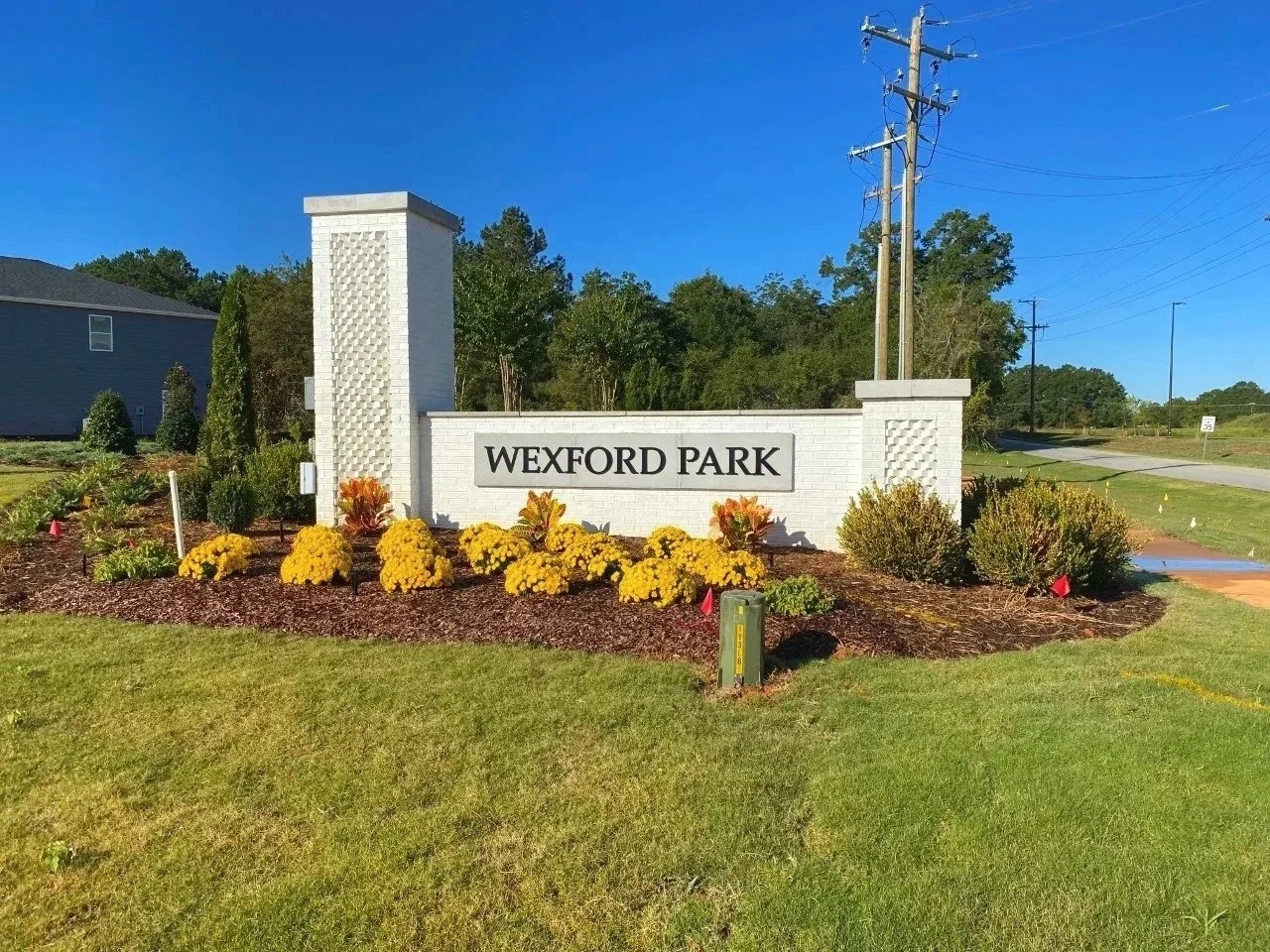 Entrance sign for Wexford Park with landscaped yellow and orange flowers, green shrubs, and trees in the background under a clear blue sky.