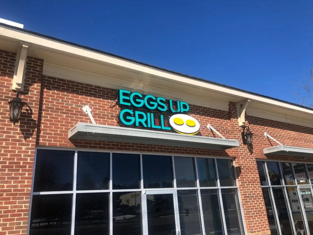 Exterior of a restaurant named 'Eggs Up Grill' with a sign displaying a smiling fried egg face, brick building, large glass windows, and outdoor wall lanterns under a clear blue sky.