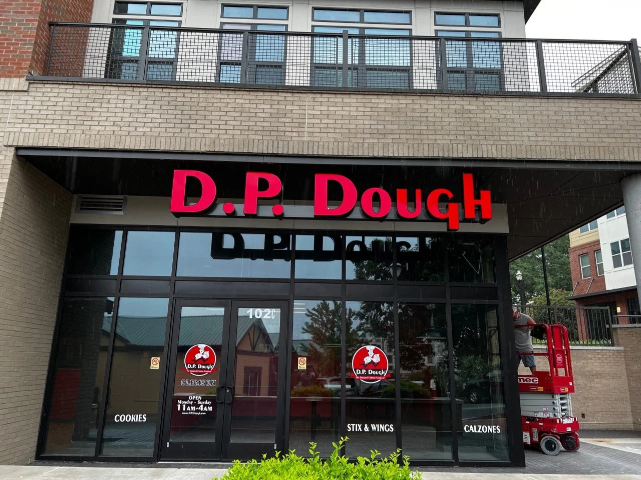 Storefront with a large sign reading 'D.P. Dough' in bold pink letters, glass doors, and windows. A worker on a red lift is visible on the right side.