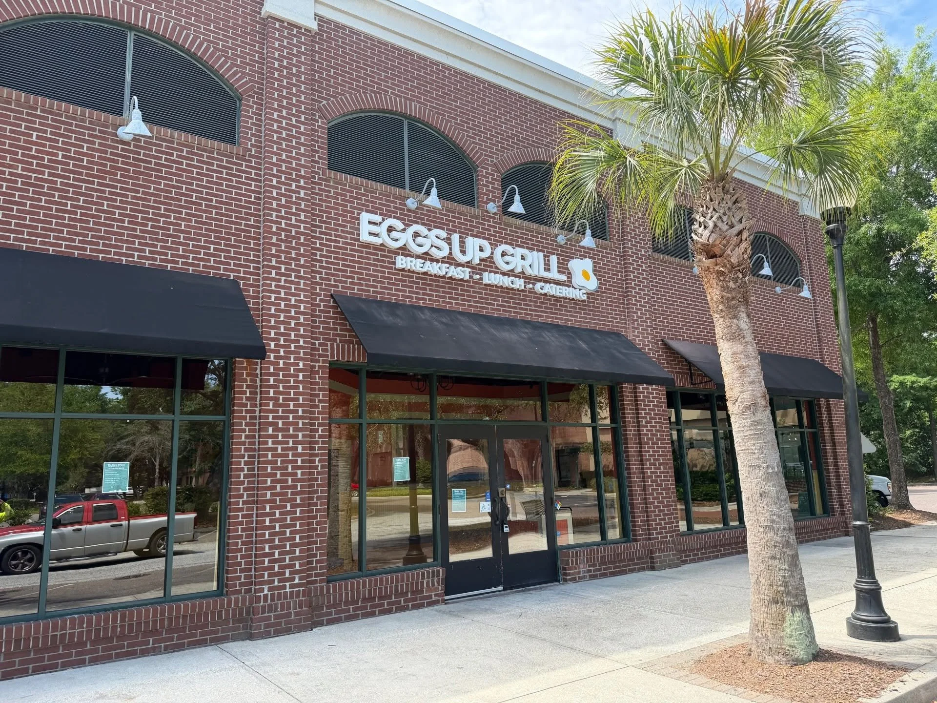Exterior of a brick building with a sign that reads "Eggs Up Grill" and lists breakfast, lunch, and catering services. There are large windows and black awnings, with a palm tree and streetlamp outside.