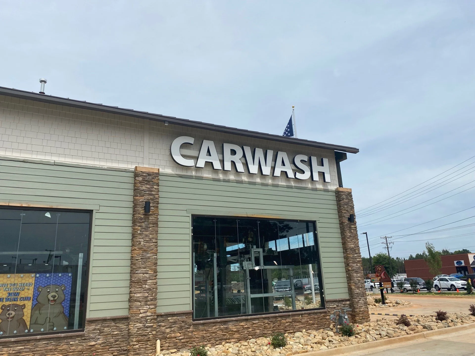 Exterior view of a car wash building with large 'CARWASH' sign, American flag on roof, large windows, and stone accents on the facade, with a parking lot and cars in the background under a cloudy sky.