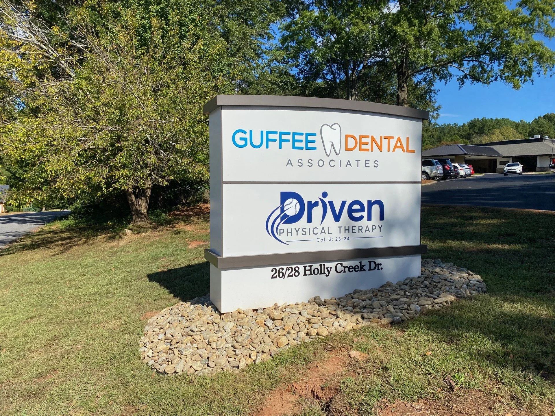 Sign for Guffee Dental Associates and Driven Physical Therapy at 26/28 Holly Creek Drive, with parked cars and a building in the background, surrounded by trees and a grassy area.