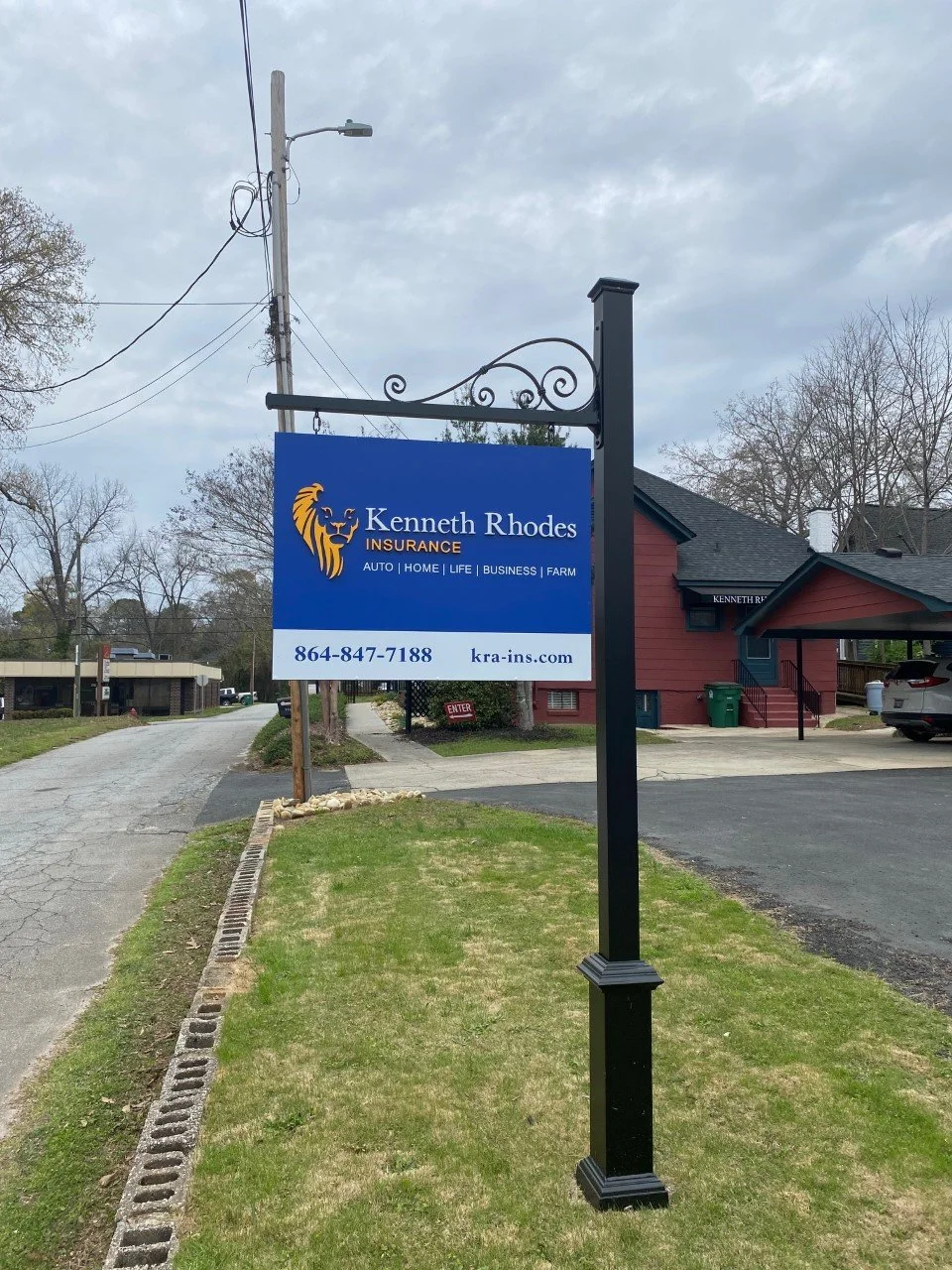 Sign for Kenneth Rhodes Insurance with lion logo and contact information, located near a road and building in a suburban area.