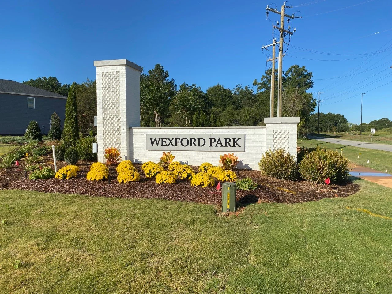 Entrance sign for Wexford Park, surrounded by yellow and orange flowers, with a white brick background and greenery, under a clear blue sky.
