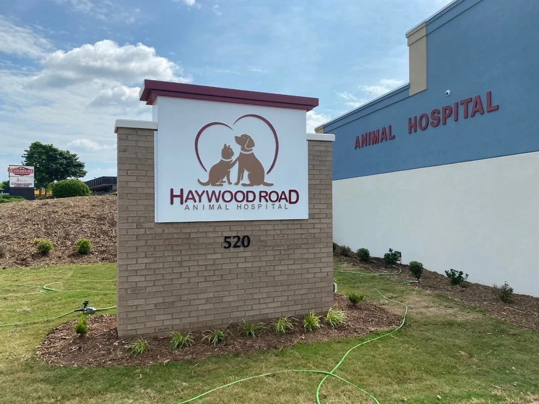 Sign for Haywood Road Animal Hospital featuring a logo with a dog and cat inside a heart, in front of a brick structure, next to a blue building with the words "Animal Hospital" on it.