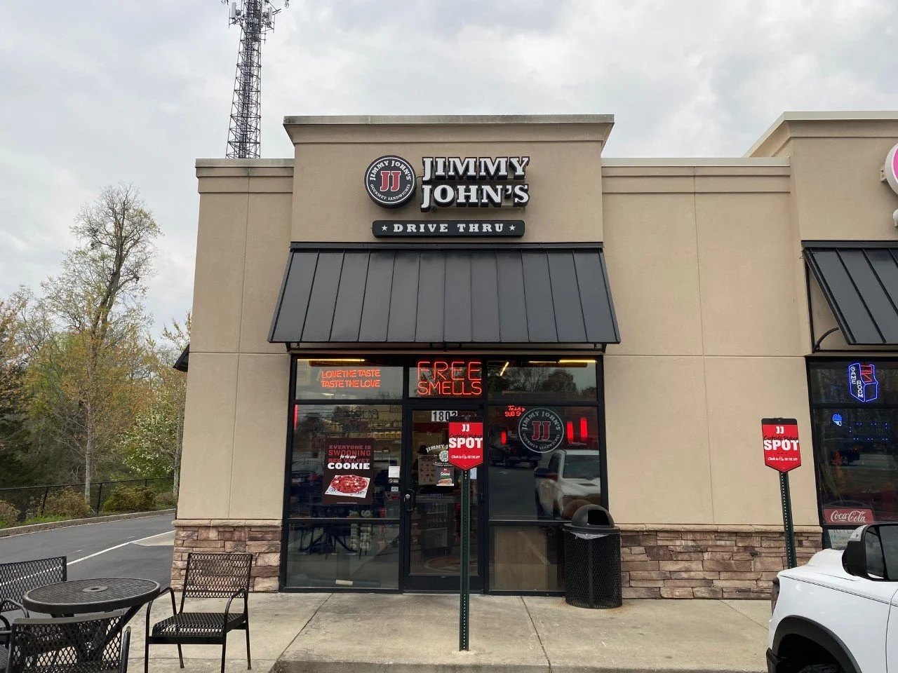 Jimmy John's restaurant storefront with a drive-thru sign, neon signs in the window, outdoor seating, and parked cars in front.