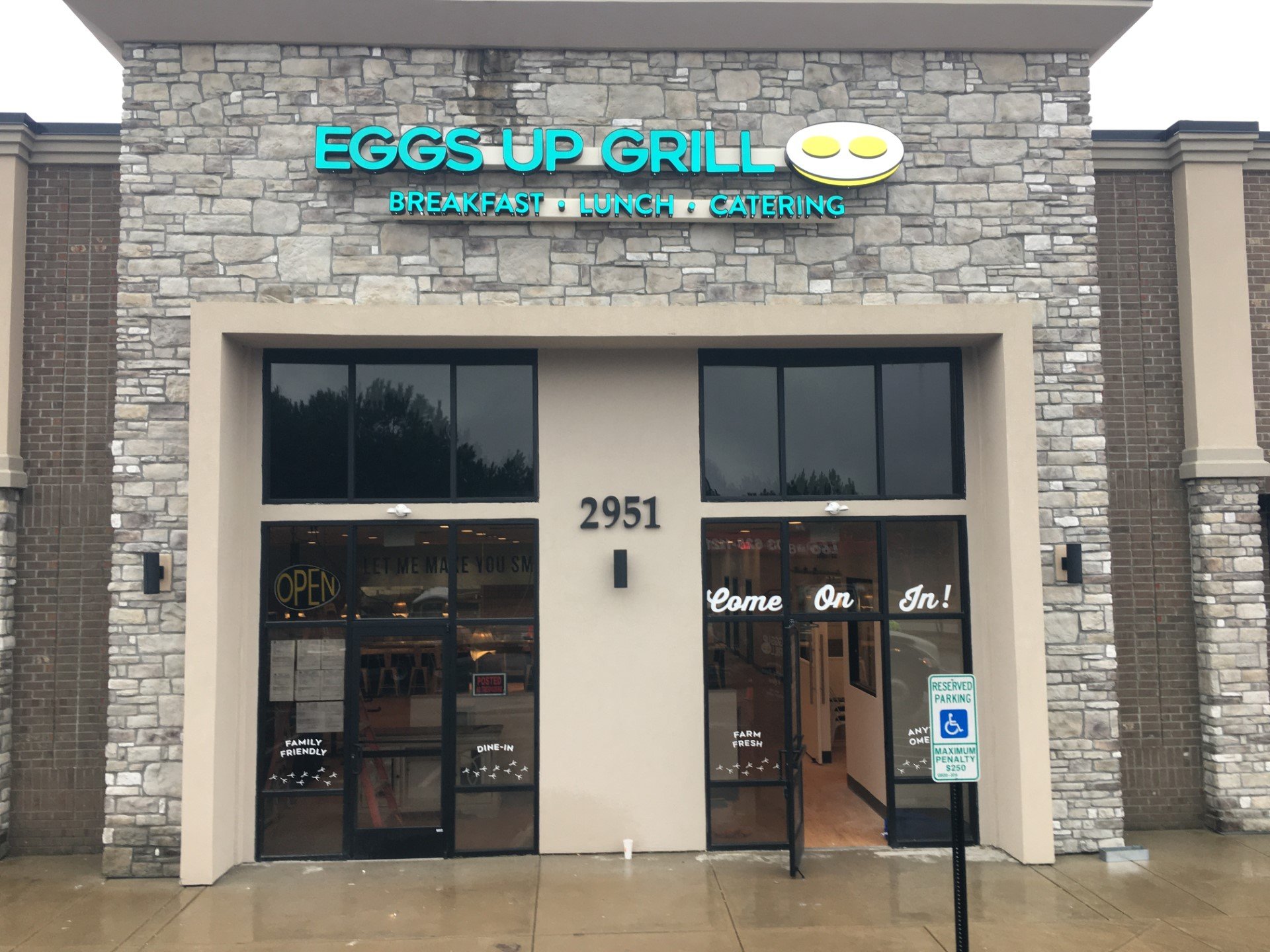 Front view of a restaurant called Eggs Up Grill with stone and brick facade, blue illuminated sign, and glass doors. Signs indicate it is open and family friendly, offering breakfast, lunch, and catering.