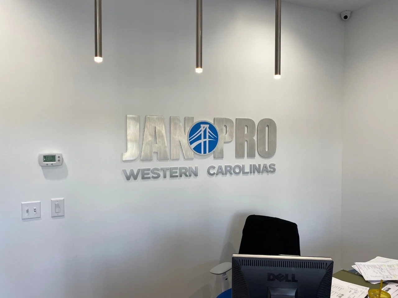 Office wall with a sign that reads 'JAKPRO Western Carolinas', a digital thermostat, light switches, and part of a desk with papers, a pen, and a Dell monitor.