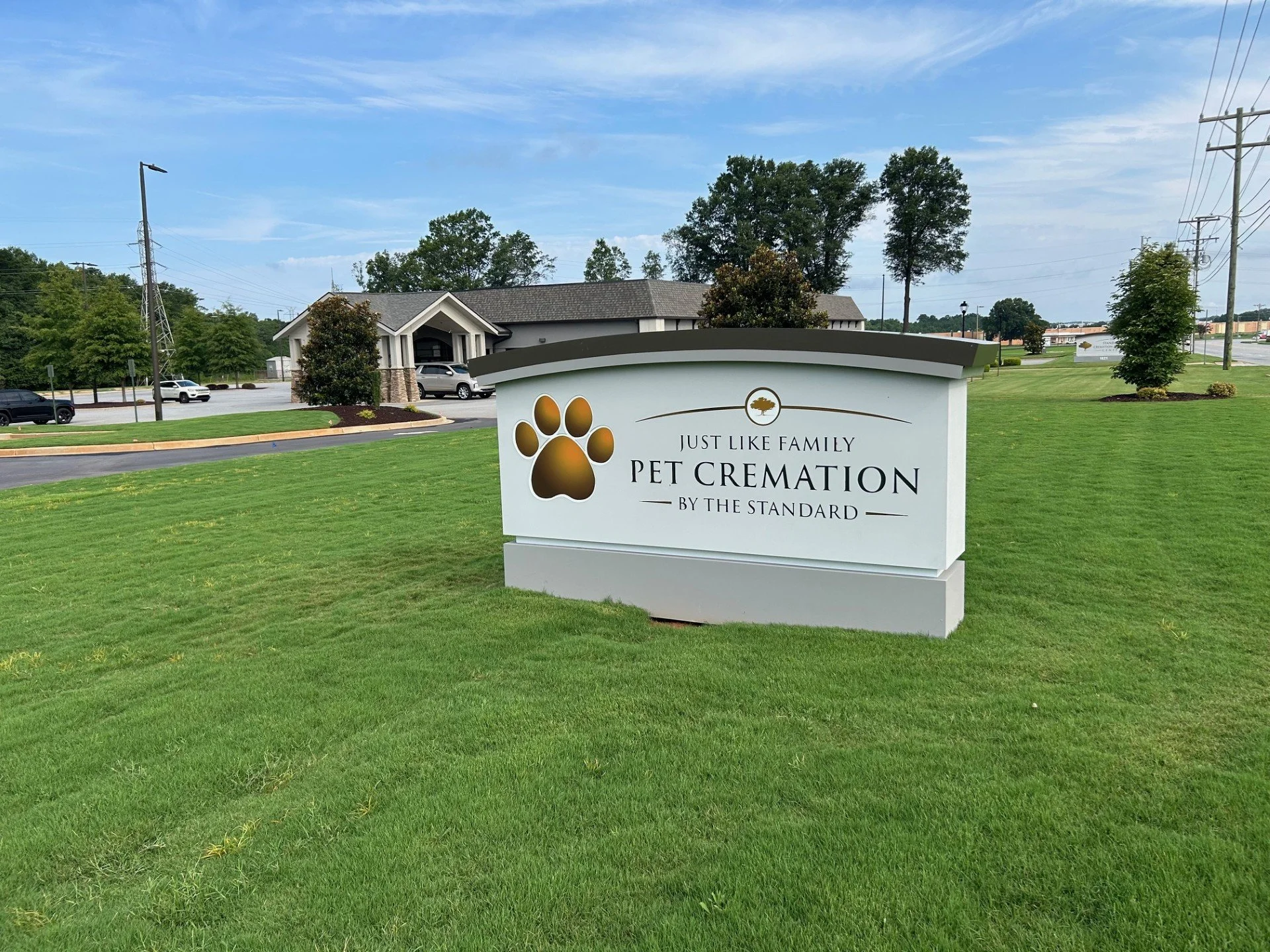 Sign for 'Just Like Family Pet Cremation' on a grassy area outside a building, with trees, parking lot, and power lines in the background.