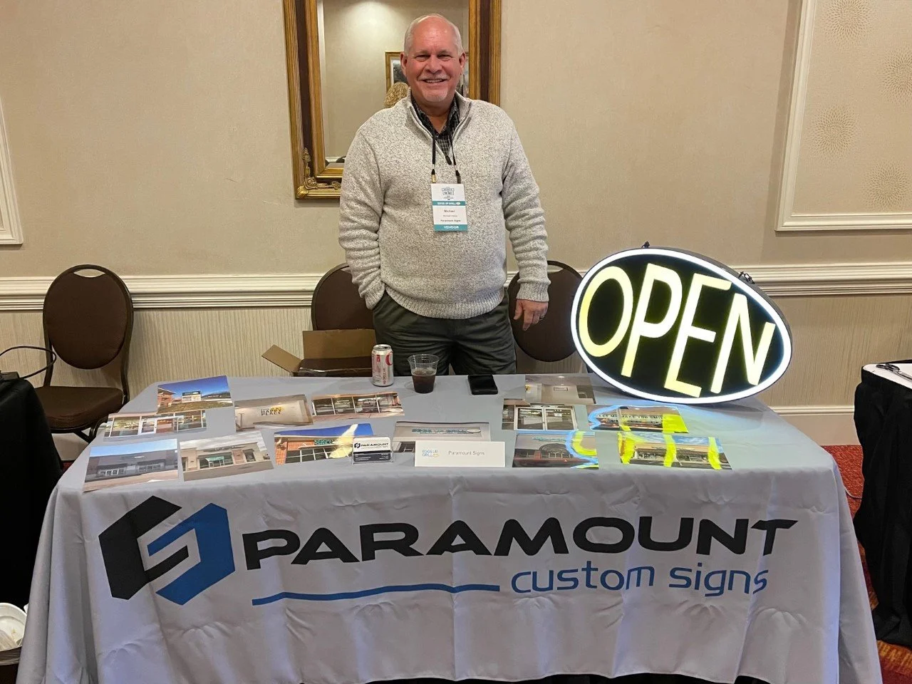 Man standing behind a table with Paramount custom signs banner, open sign, and promotional materials at a trade show.